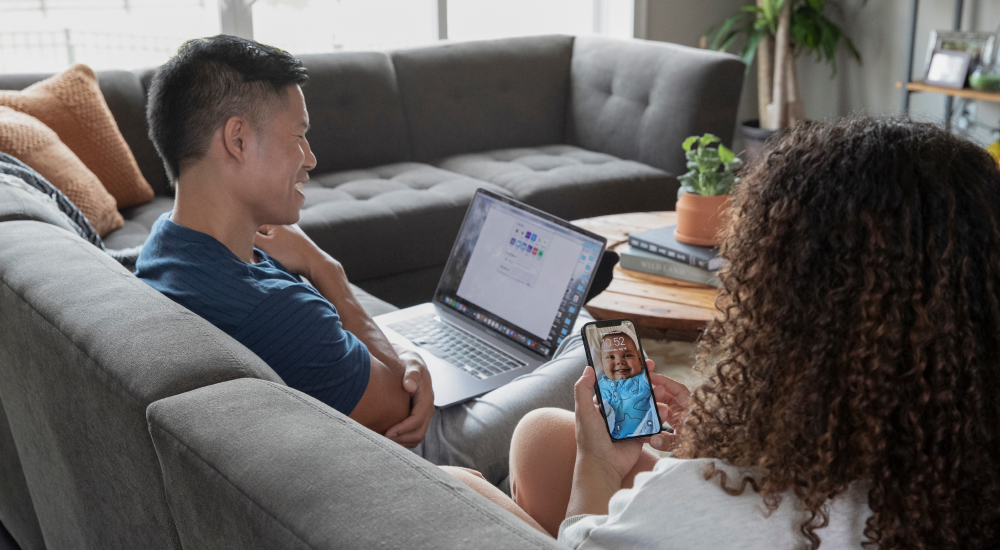 A couple sitting on a couch. The man has his laptop on his lap and the woman is looking at her mobile phone that has a background screen of a baby.