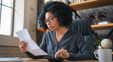 A woman is sitting in an office. She's holding a paper document in one hand and working on a calculator with the other.